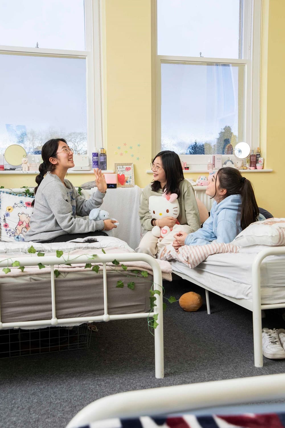 Three people sitting on beds in a cozy room, smiling and chatting. One holds a stuffed toy. The room has two large windows and decorative items.