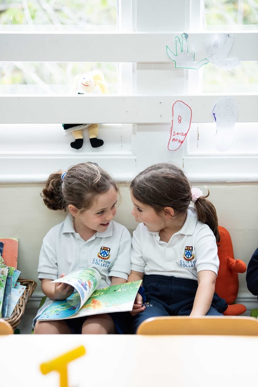 Two young girls in school uniforms sit by a window, sharing a colorful book and smiling at each other.