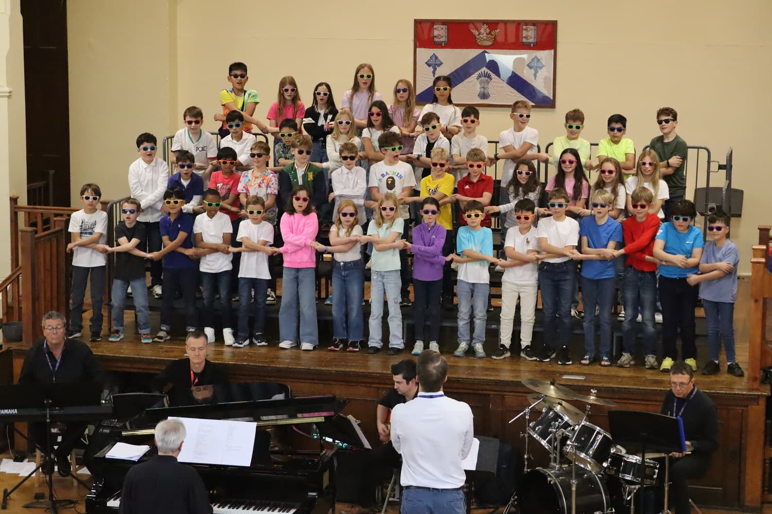 A group of children wearing sunglasses perform on stage, with musicians playing instruments in front of them.