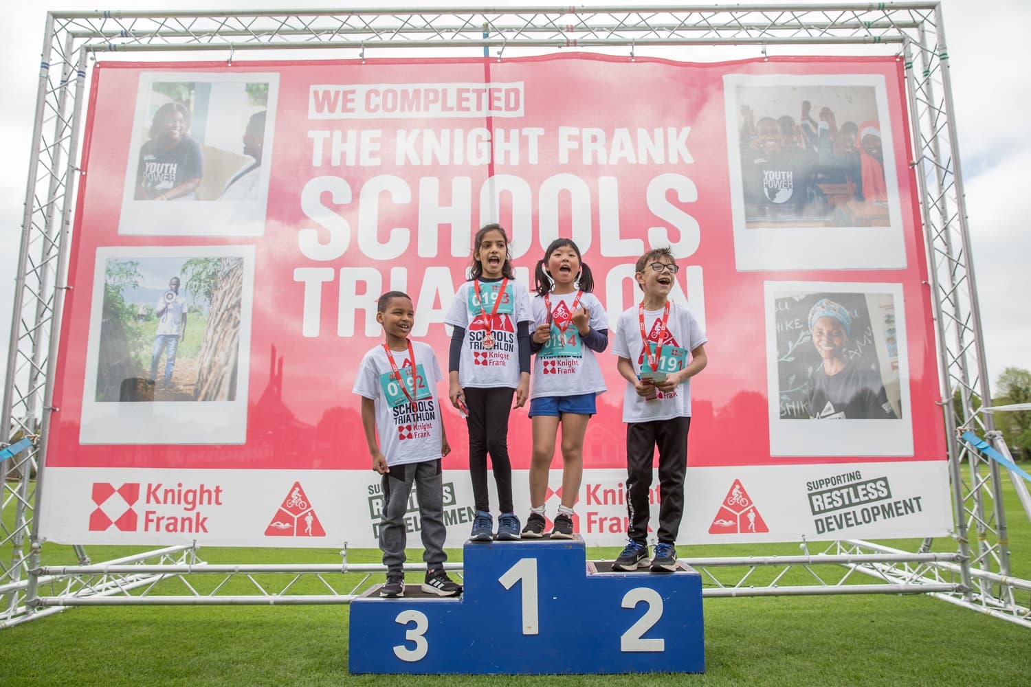 Four children stand on a winners' podium, wearing medals and race numbers, in front of a large "Knight Frank Schools Triathlon" banner.