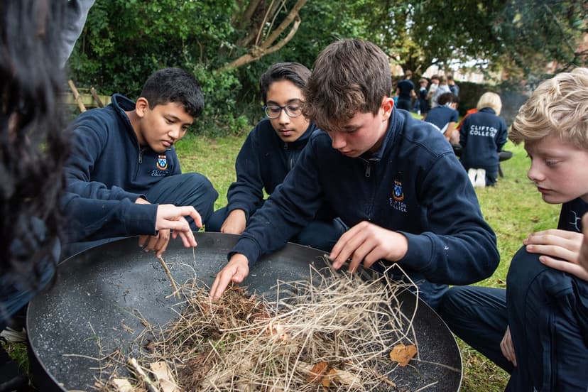 Students in uniforms gather outdoors, focusing on starting a fire with kindling in a large metal bowl, surrounded by greenery.