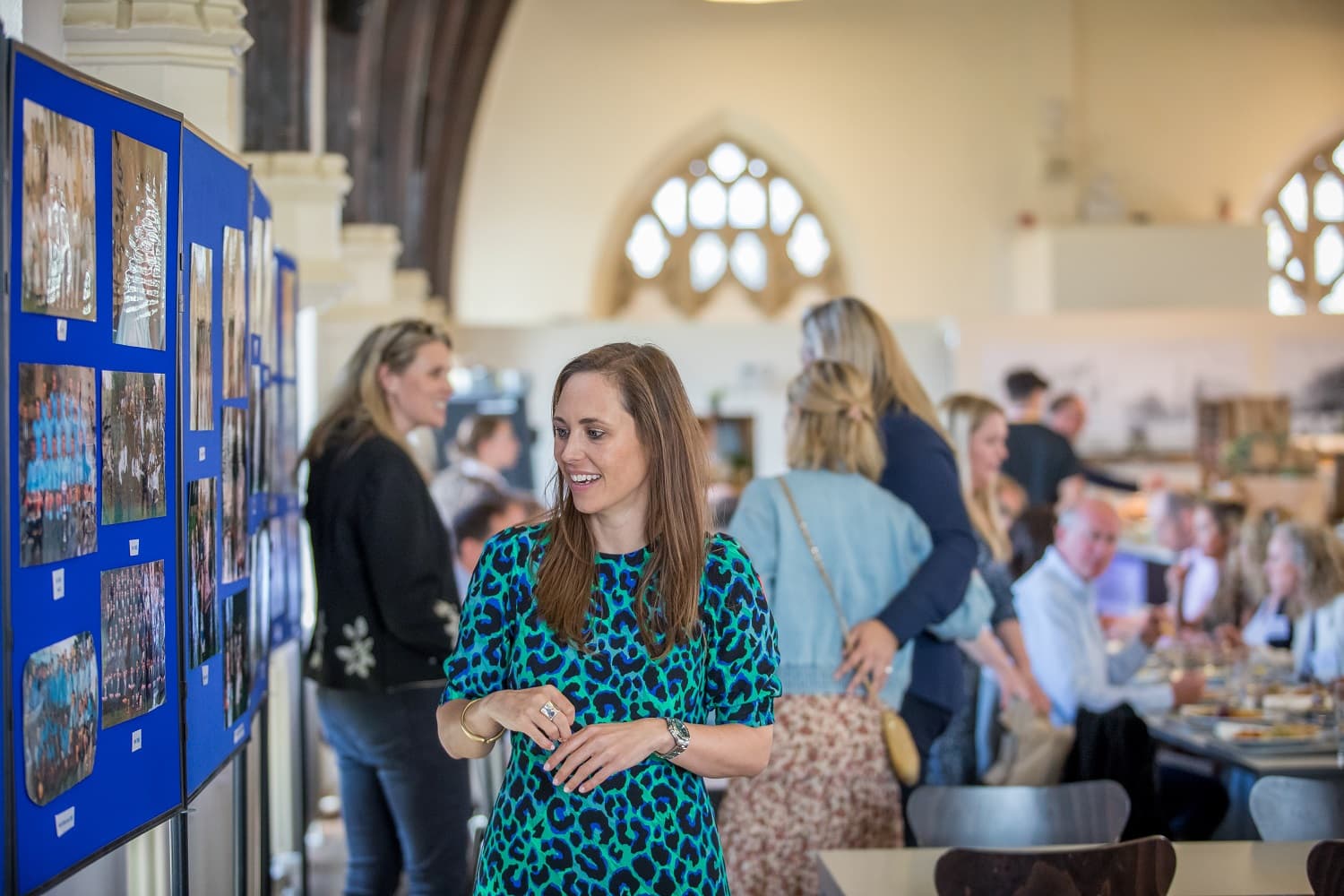 A woman in a blue patterned dress looks at photos on a display board in a busy room with people socializing in the background.
