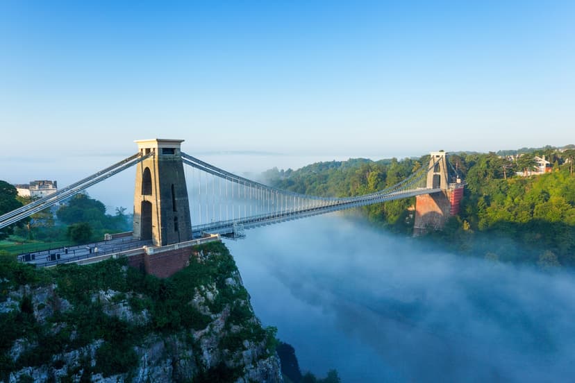 Suspension bridge spanning a misty river gorge, surrounded by lush greenery under a clear blue sky.