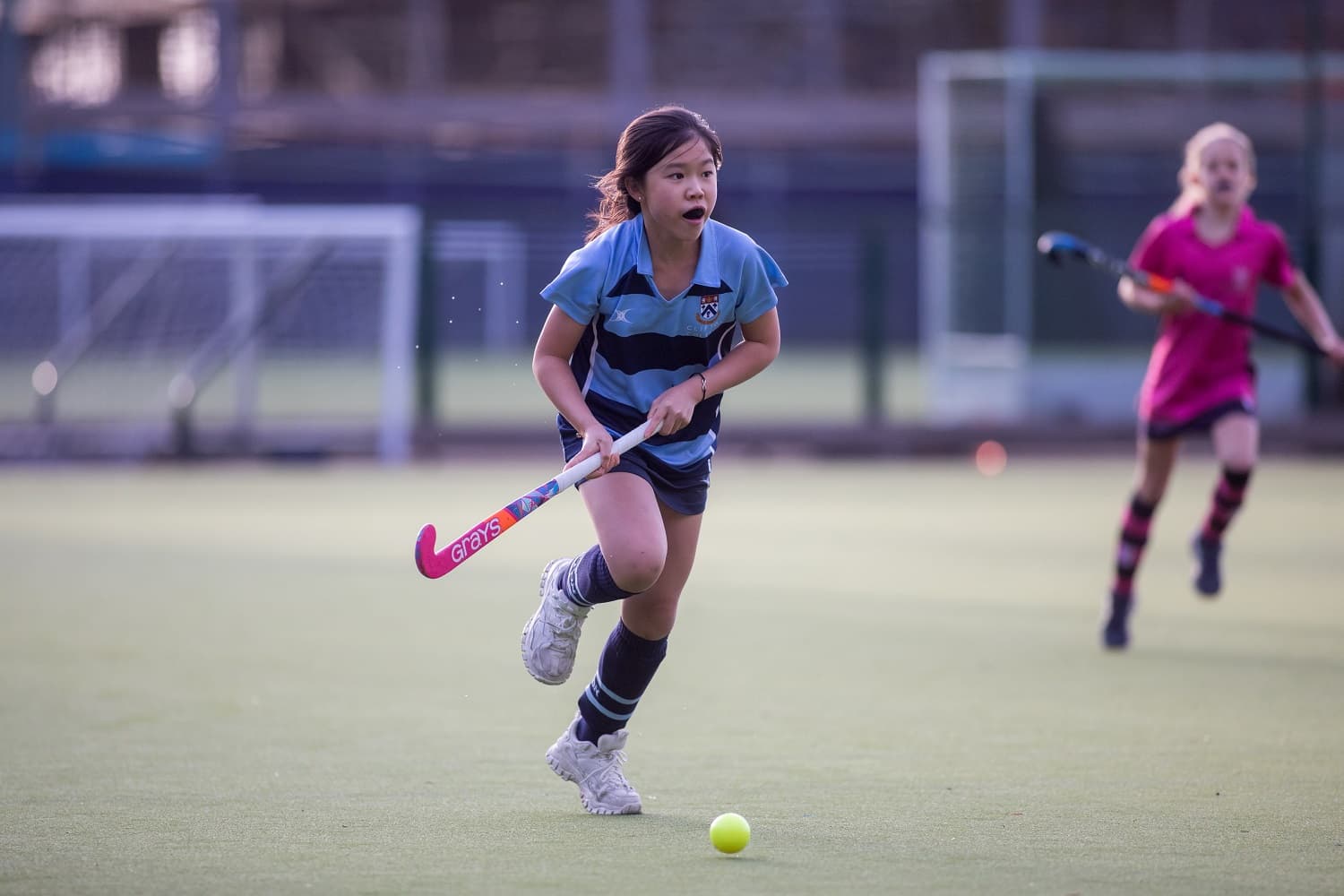 A young girl in a blue and black uniform plays field hockey, running with the ball on a grassy field. Another player is visible in the background.