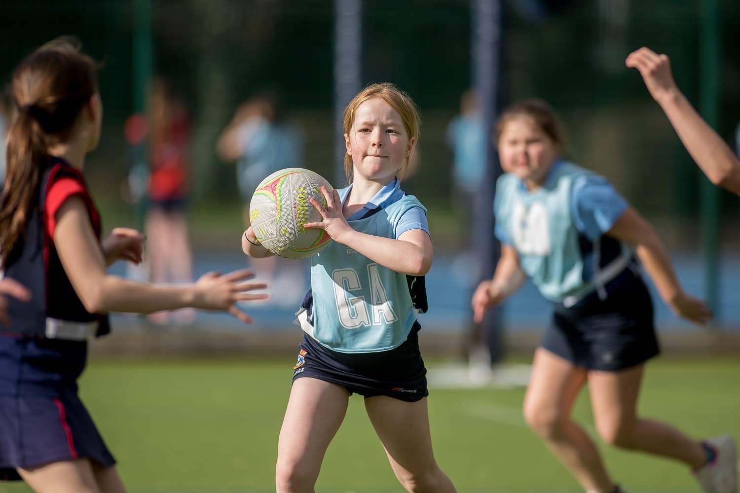 Girls playing netball outdoors, with one girl in focus catching the ball. They are wearing sports uniforms on a sunny day.
