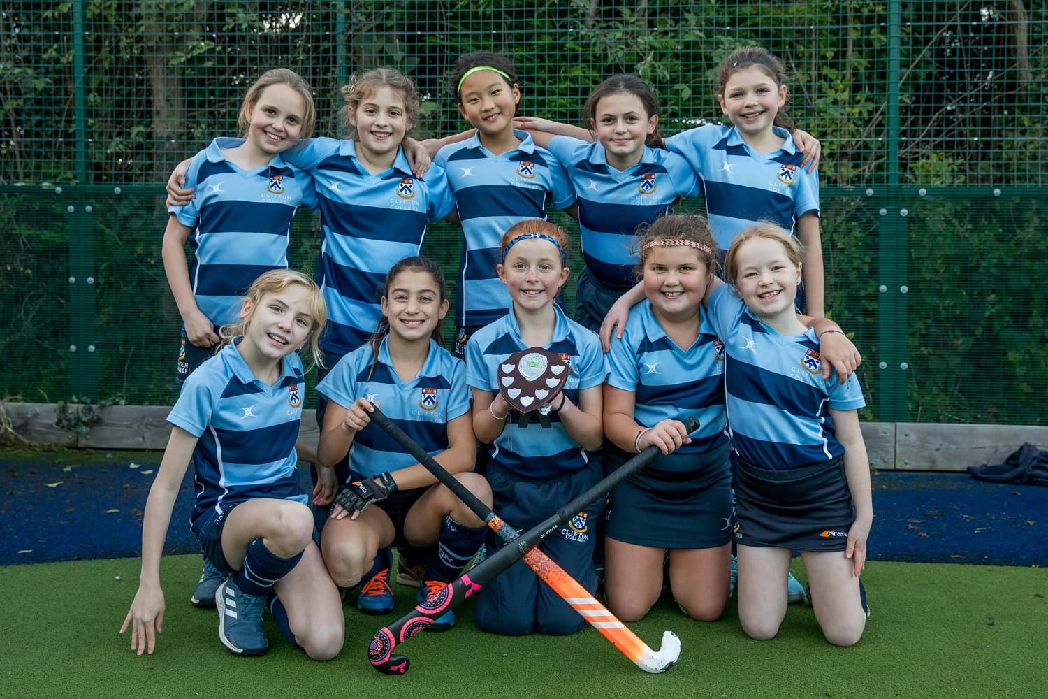 A girls' field hockey team in blue uniforms poses with a trophy on a turf field, smiling and celebrating their victory.