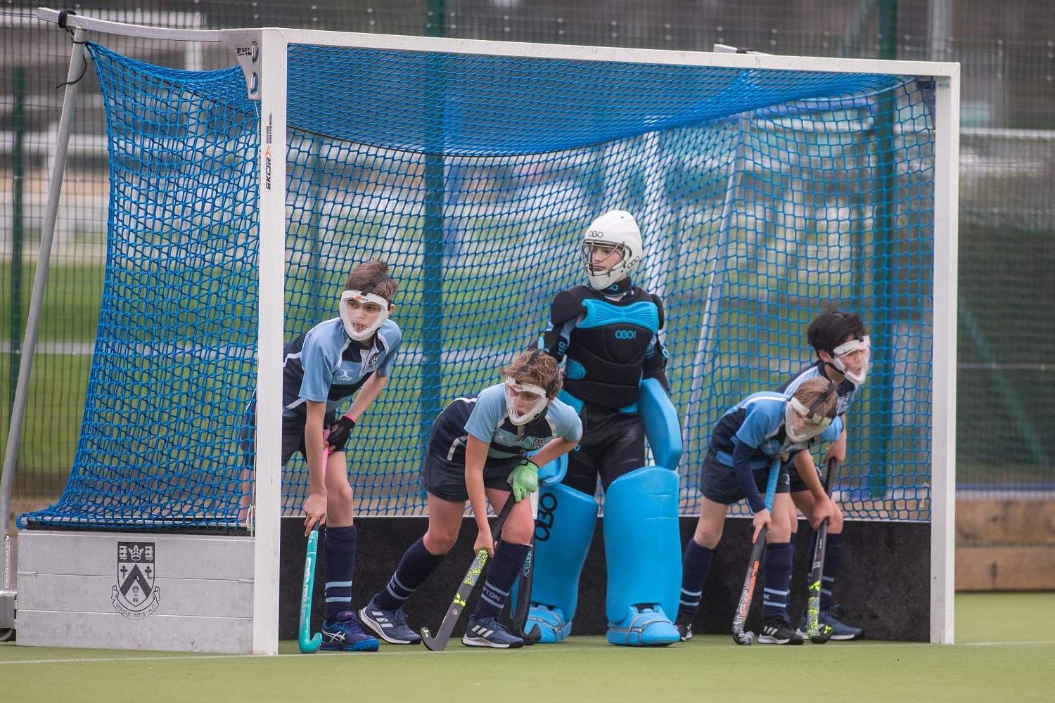 Four young field hockey players and a goalie in blue uniforms and protective gear stand ready in front of a goal on a turf field.