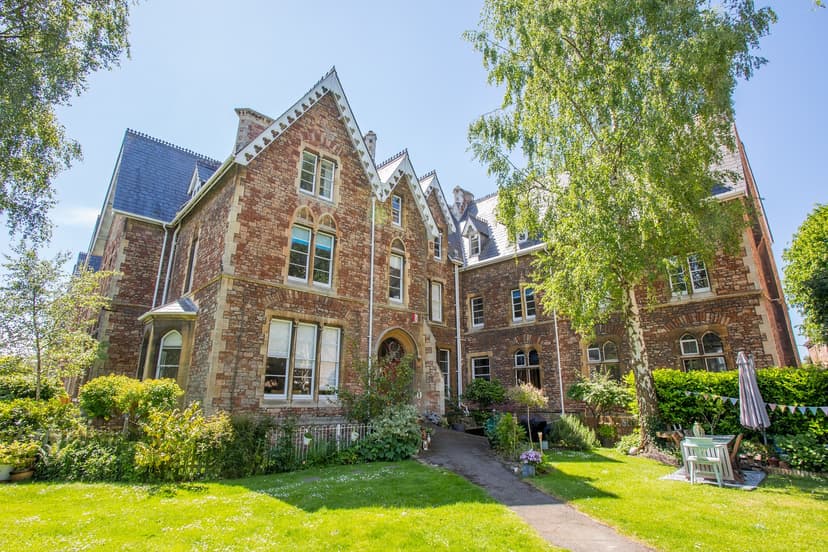 Historic brick building with arched windows, surrounded by lush greenery and a garden path under a clear blue sky.