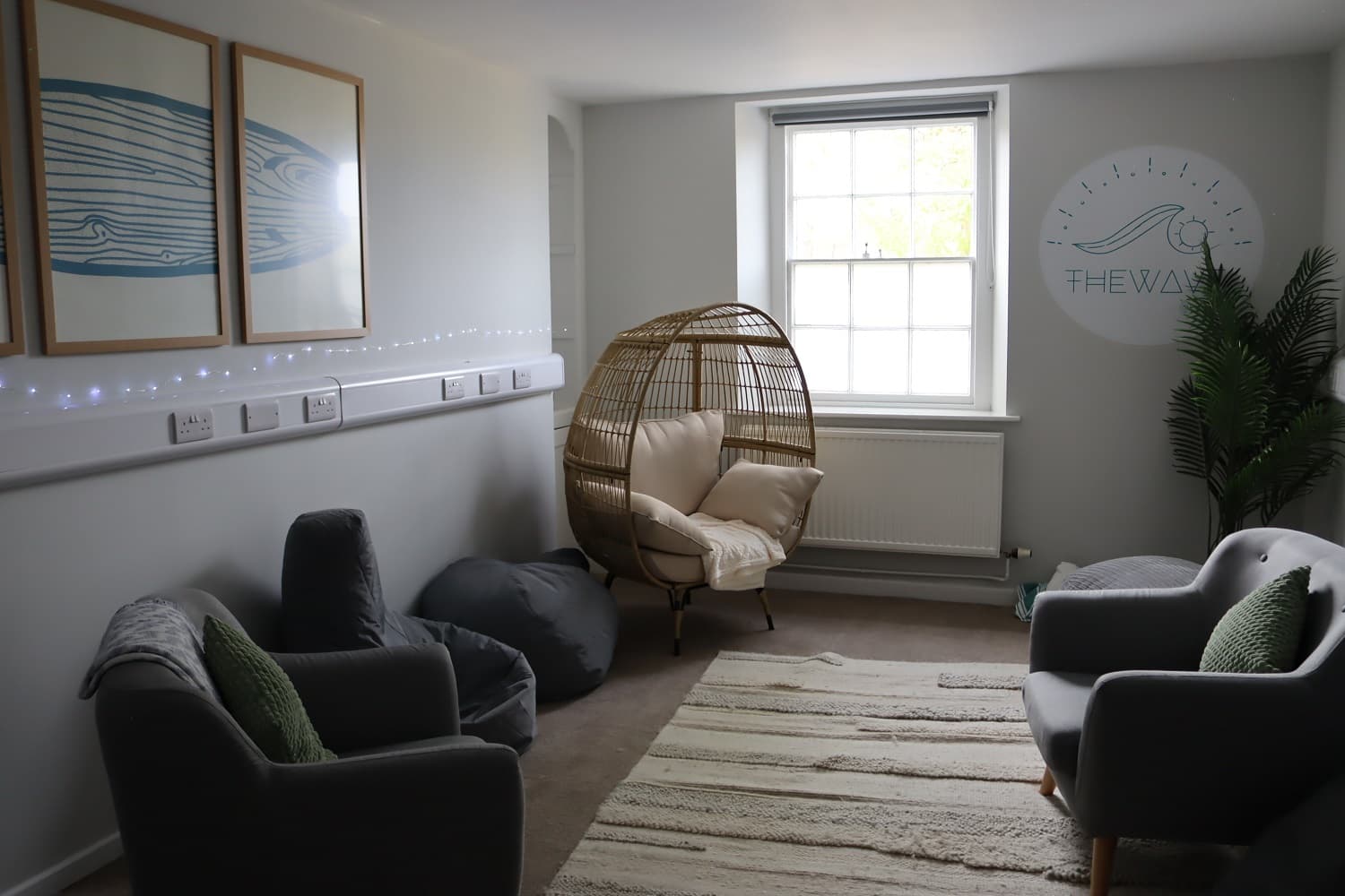 Cozy room with two gray chairs, a wicker egg chair, bean bag, framed art, and a potted plant. Soft lighting and a window brighten the space.