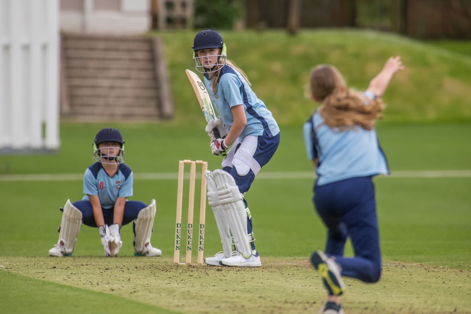 Young cricketer batting, another bowling, and a wicketkeeper crouching on a grassy field. All are wearing blue uniforms and helmets.