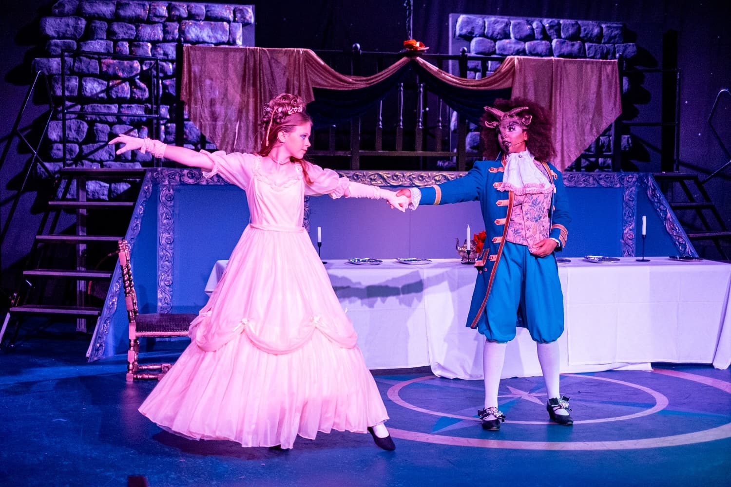 Two performers in period costumes dance on stage; one in a yellow gown, the other in a blue coat with a beast-like mask.