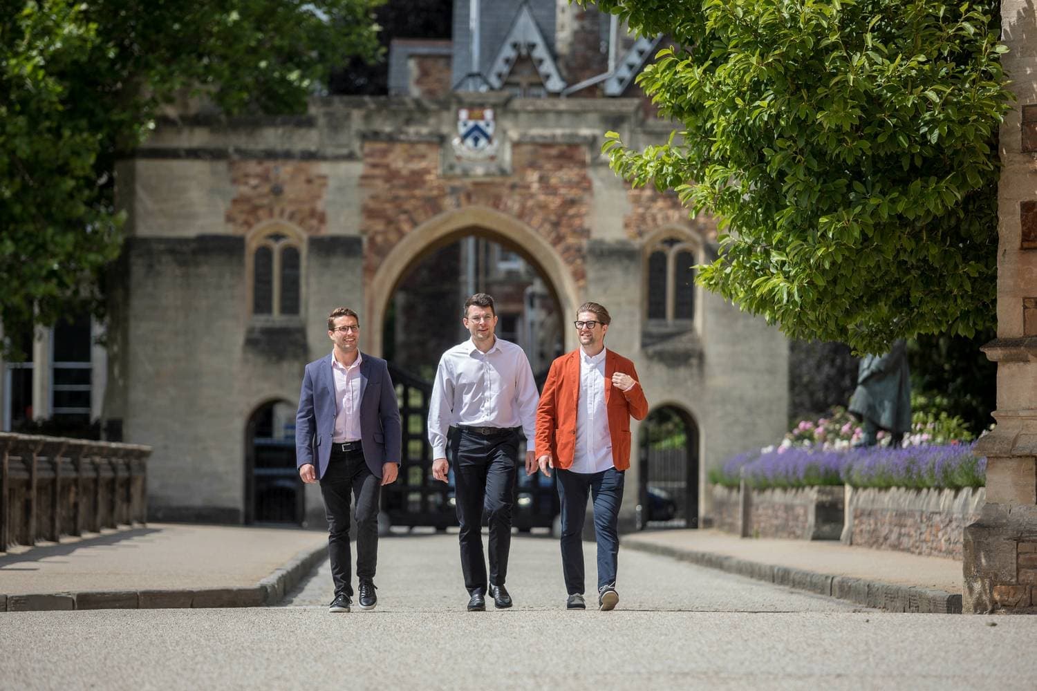 Three men walking on a path towards a historic stone building with arched windows, surrounded by greenery.