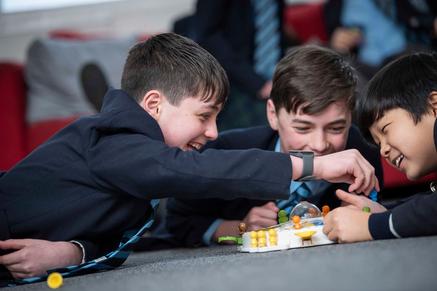 Three boys in school uniforms playing a board game on the floor, smiling and focused on the game pieces.