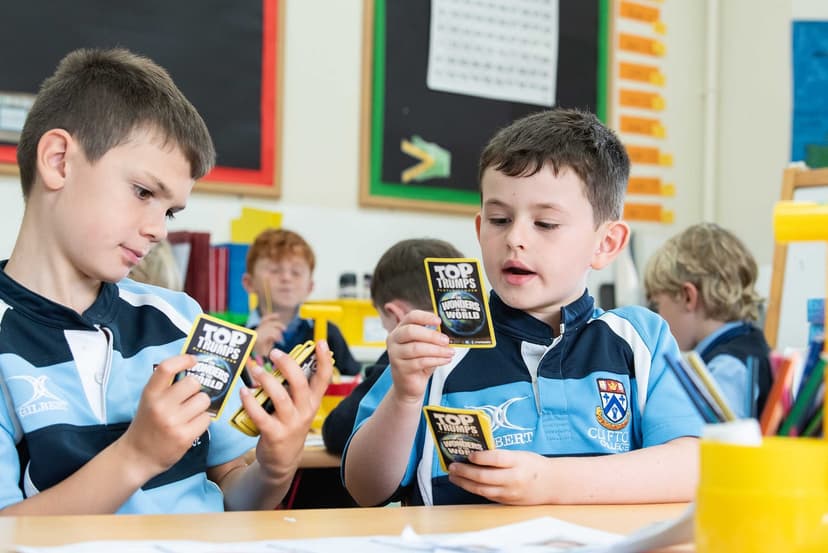 Two boys in school uniforms sit at a desk playing with "Top Trumps" cards. Other children are visible in the background.