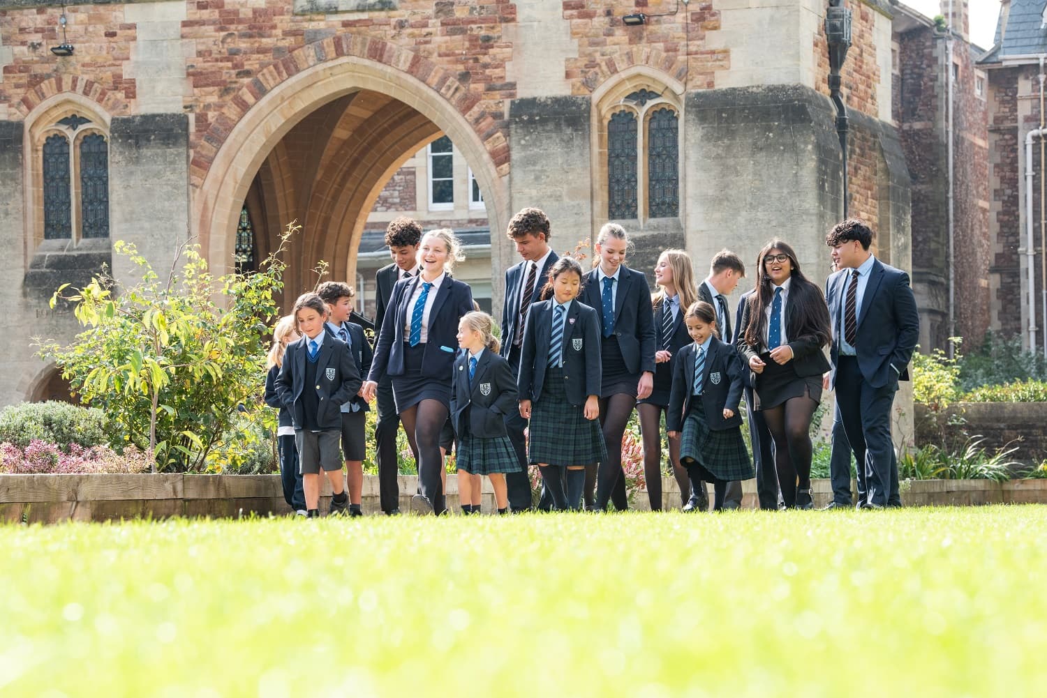 A group of students in uniforms walking on a grassy area in front of a historic building with arched windows.