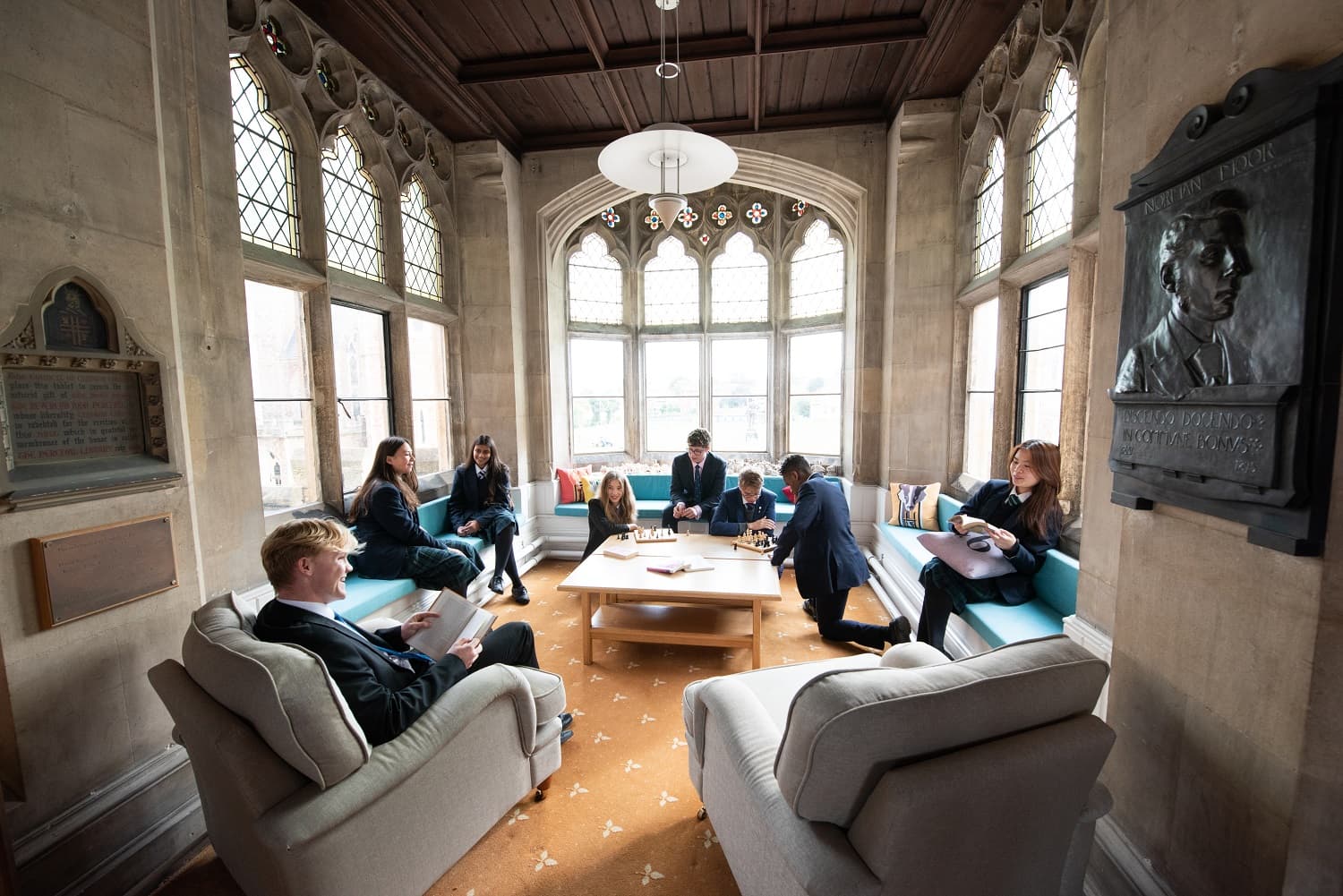 A group of people in formal attire sit and converse in an elegant room with arched windows and a high ceiling.