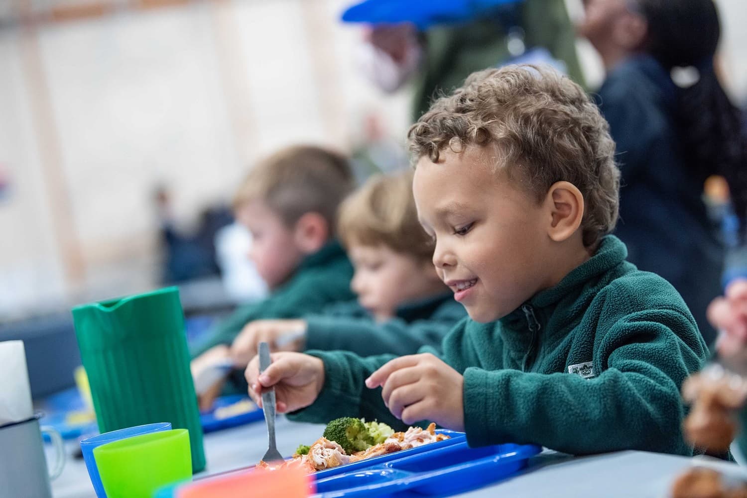 Young boy in a green sweater eating lunch at a cafeteria table, surrounded by other children with colorful trays and cups.