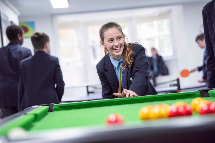 A smiling student in a school uniform plays pool, surrounded by peers in a bright room.