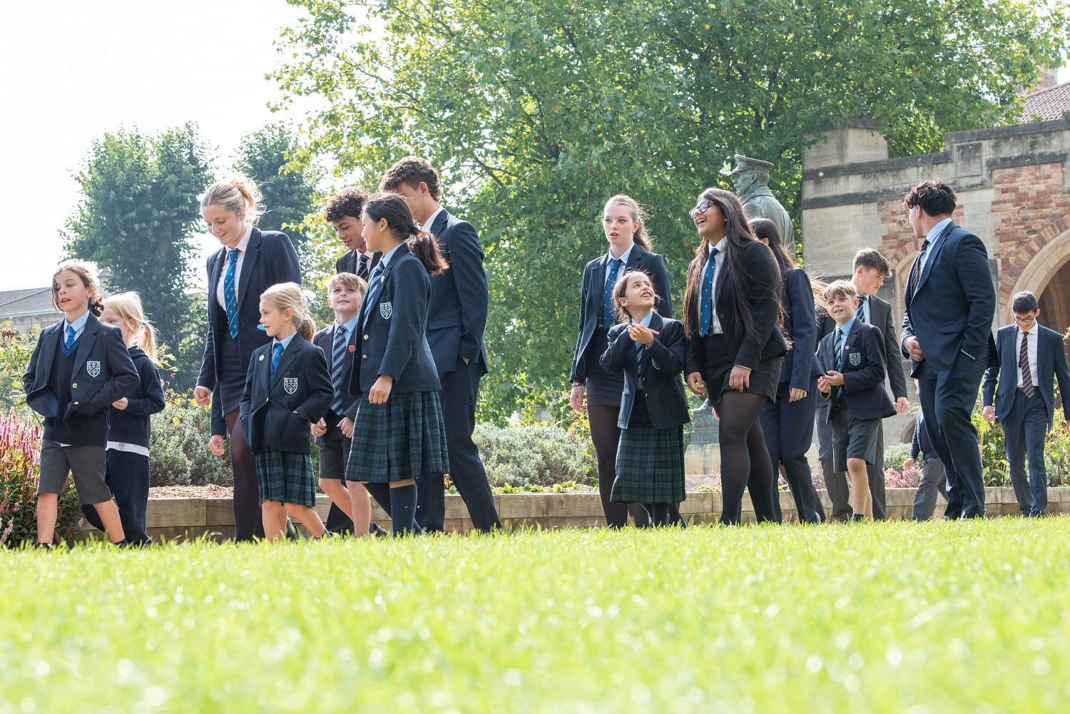 A group of schoolchildren in uniforms walking outdoors on a sunny day, with trees and a building in the background.