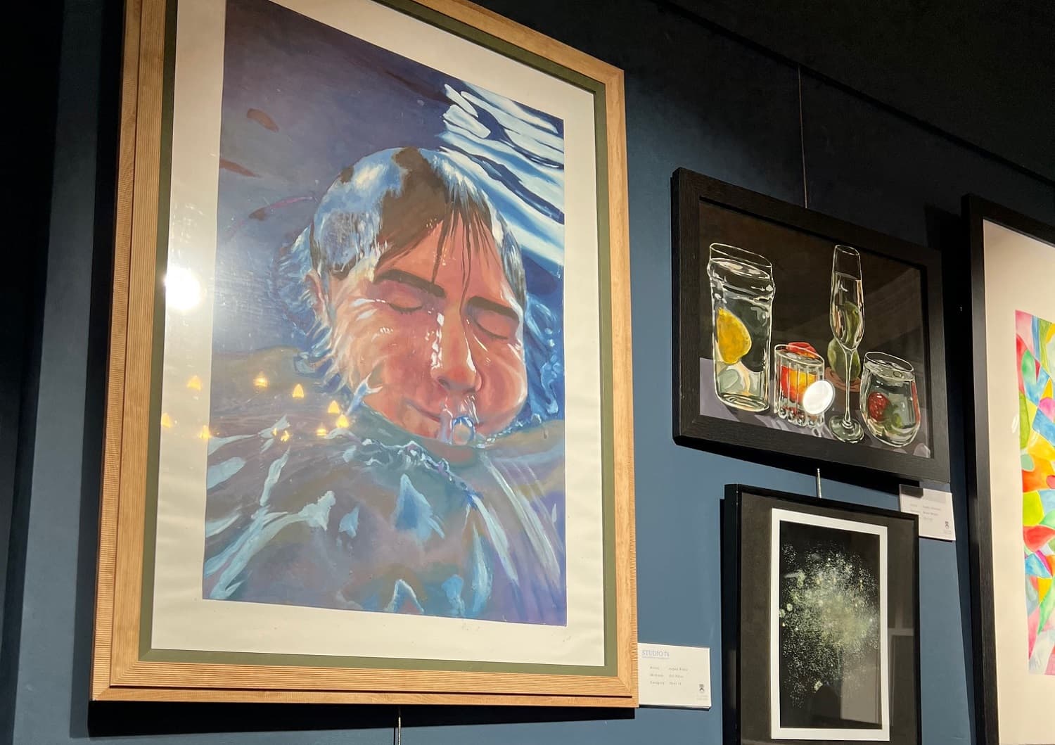 A gallery wall displaying various artworks, including a painting of a person underwater, a still life with jars, and an abstract piece.