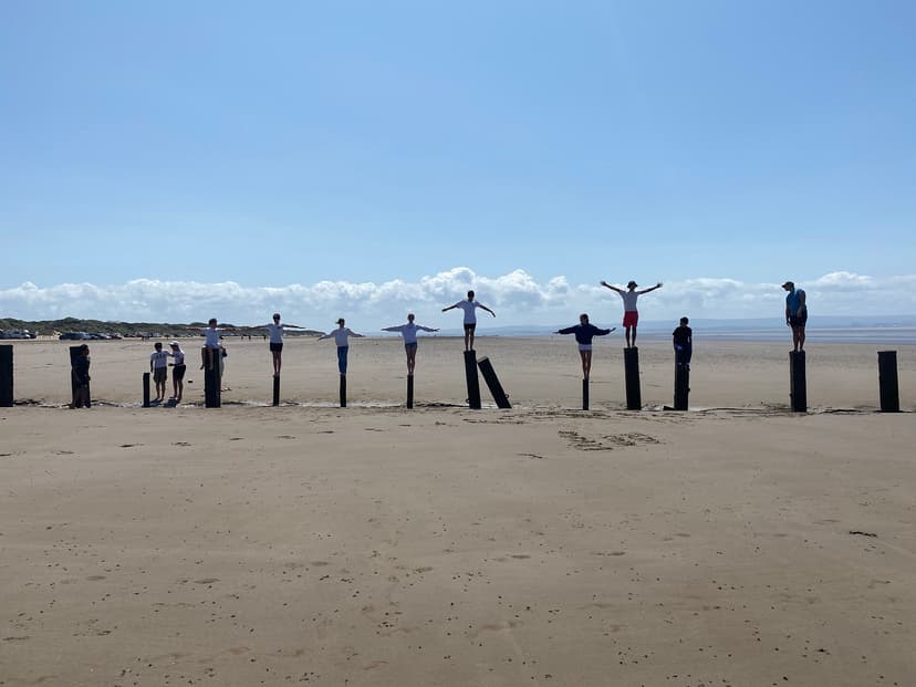 People balancing on wooden posts at a sandy beach, with clear skies and distant hills in the background.
