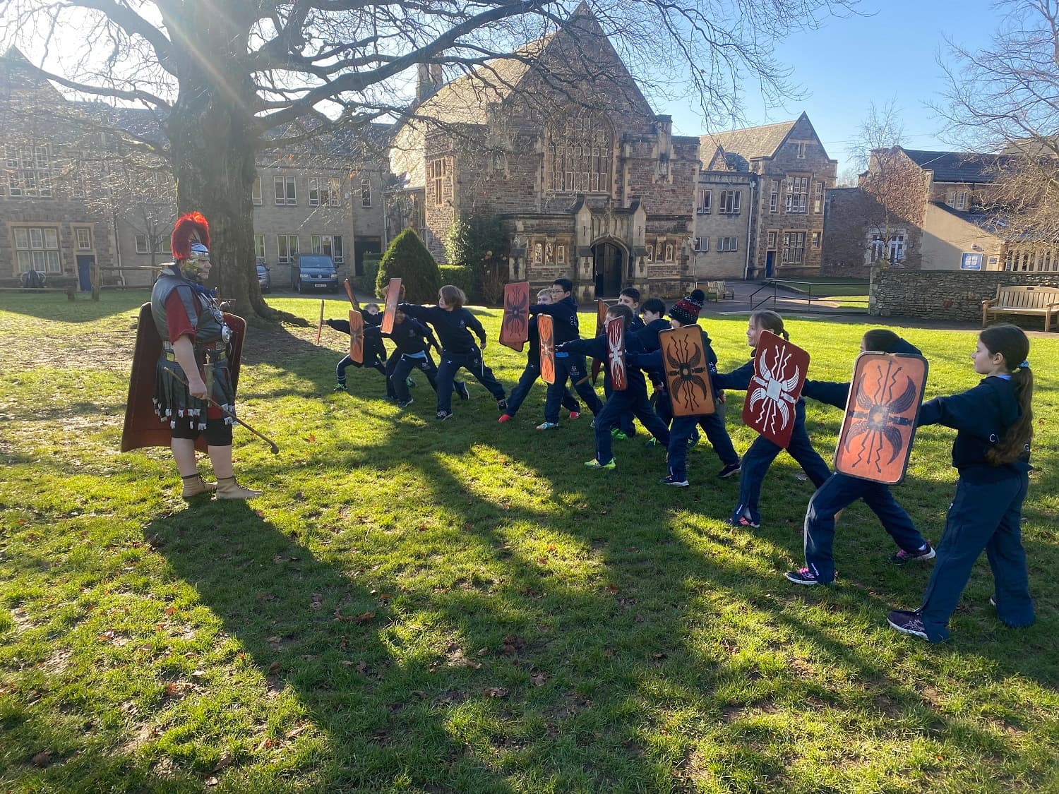 A person in Roman soldier attire instructs a group of children holding shields in a grassy area near historic buildings.