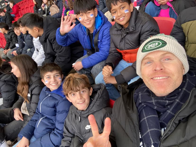 A group of people, including children and an adult, smiling and wearing warm clothing, sit in stadium seats. The adult holds up a peace sign.