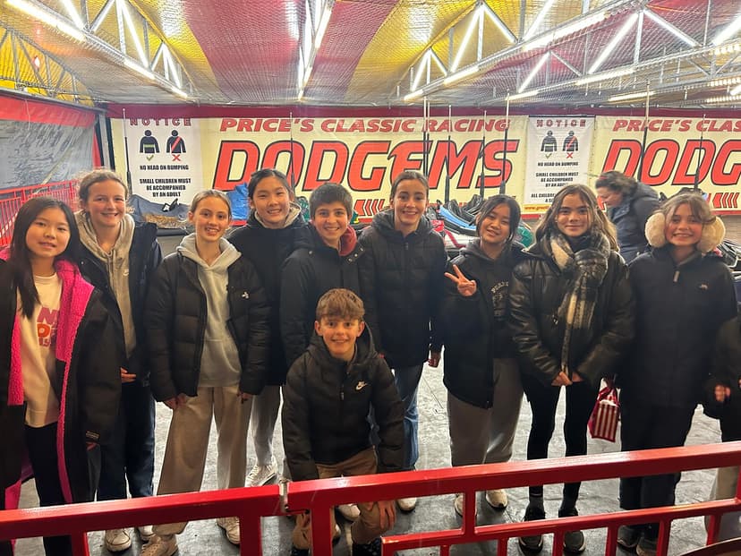 Group of smiling people in winter clothing standing in front of a "Dodgems" ride at an amusement park.