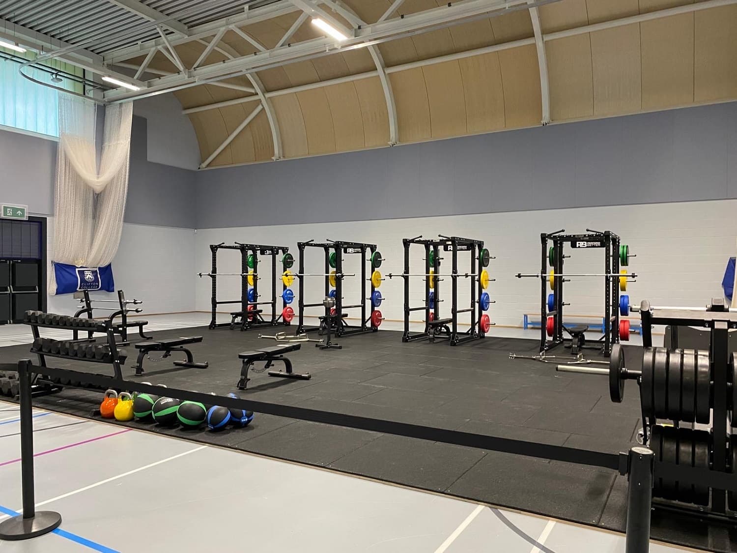 A gym with multiple squat racks, colorful weight plates, benches, kettlebells, and medicine balls on a rubber floor.