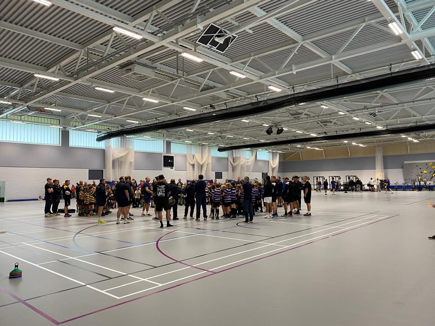 A large group of people gathers in a spacious indoor sports hall with high ceilings and bright lighting.