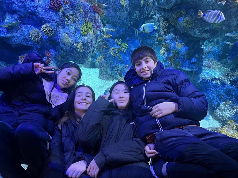 Four people in winter jackets pose happily in front of an aquarium tank, surrounded by colorful fish and coral.