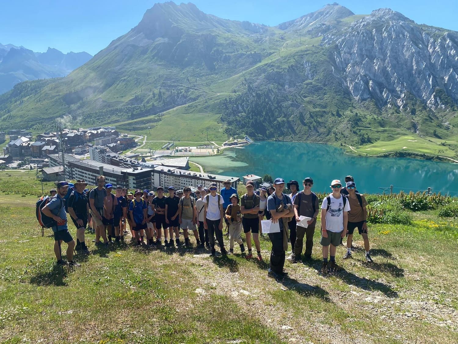 A group of hikers stands on a grassy hill with a scenic backdrop of mountains, a lake, and a village under a clear blue sky.
