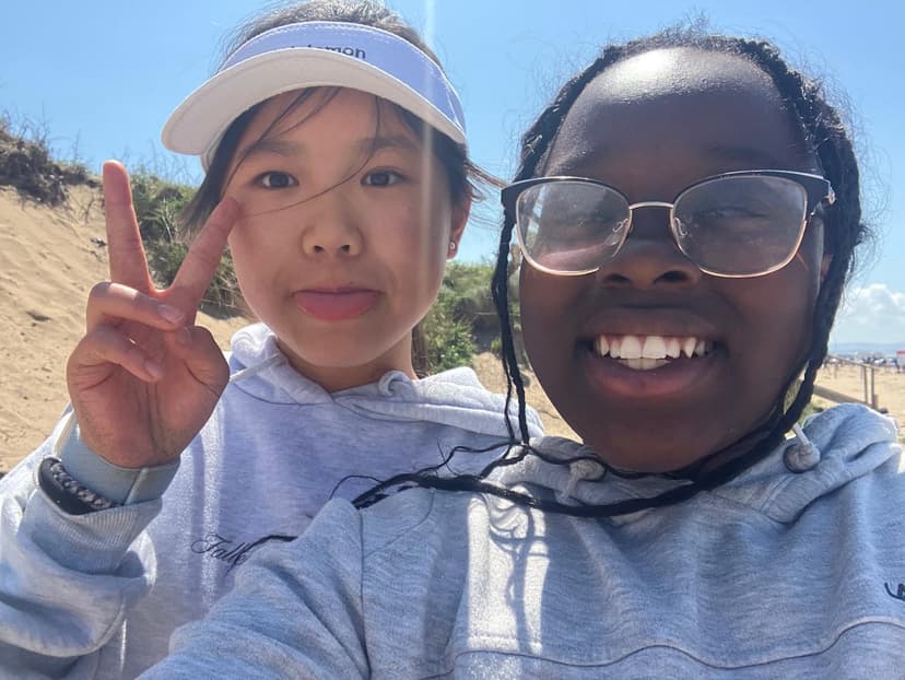 Two friends smiling at the beach; one making a peace sign and the other wearing glasses. Both in hoodies, with sand and sky in the background.