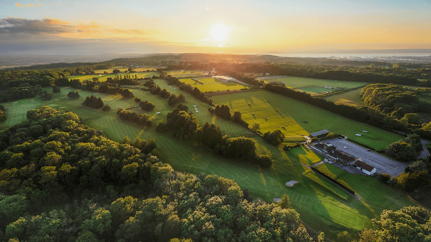 Aerial view of a golf course at sunset, with lush green fairways, surrounding trees, and a clubhouse, under a vibrant sky.