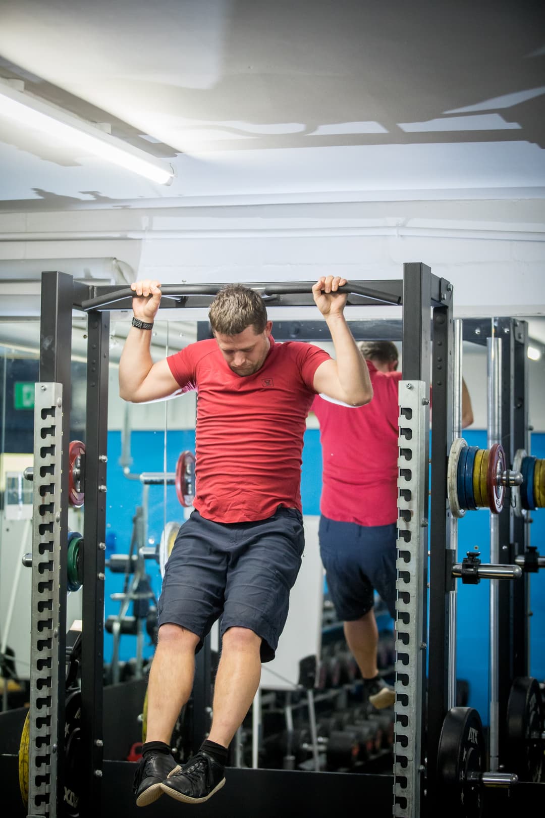 Man in a red shirt and black shorts performing pull-ups in a gym, with weights and mirrors in the background.