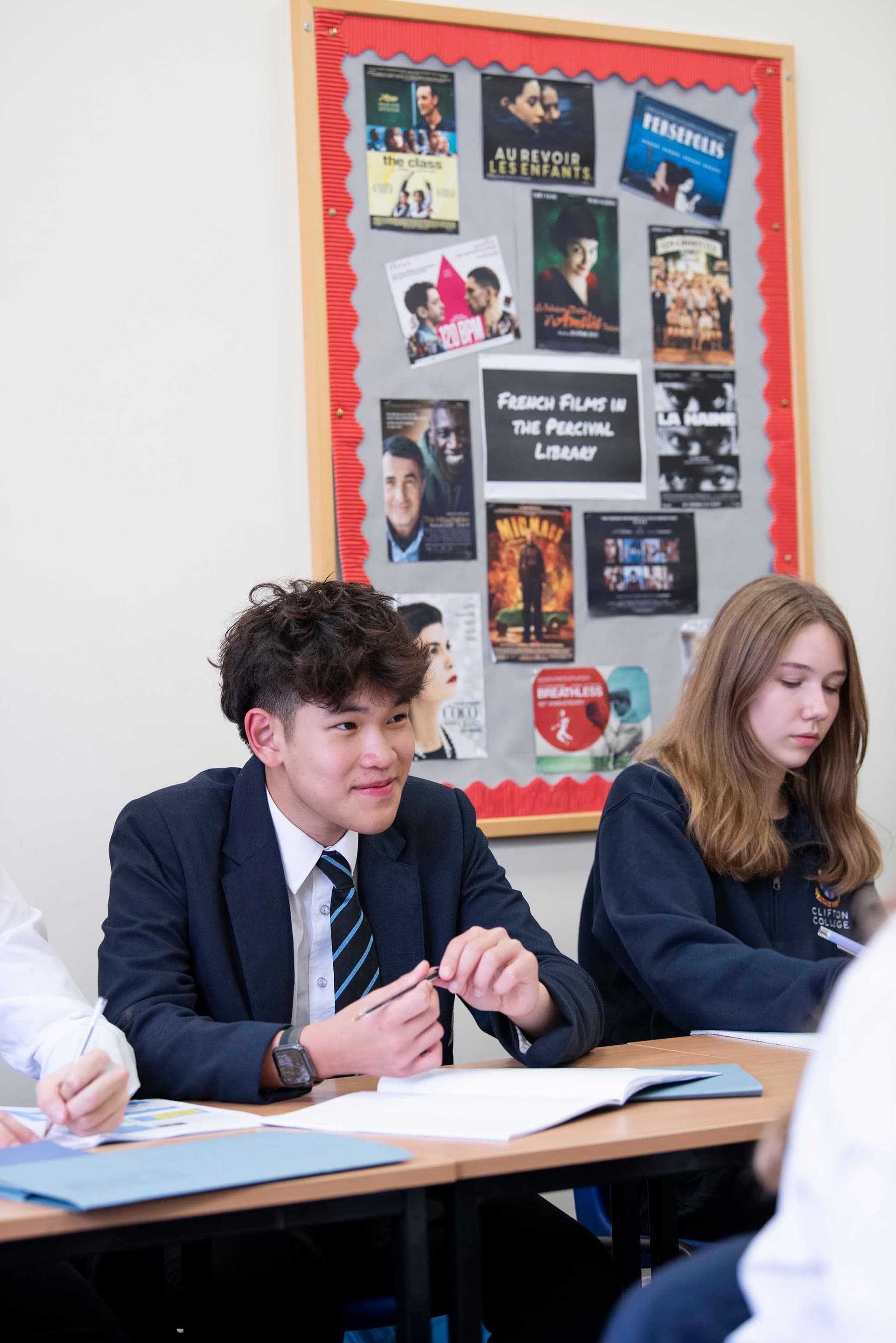 Students in a classroom, one wearing a blazer and tie, another in a sweatshirt, with a bulletin board featuring French film posters behind them.