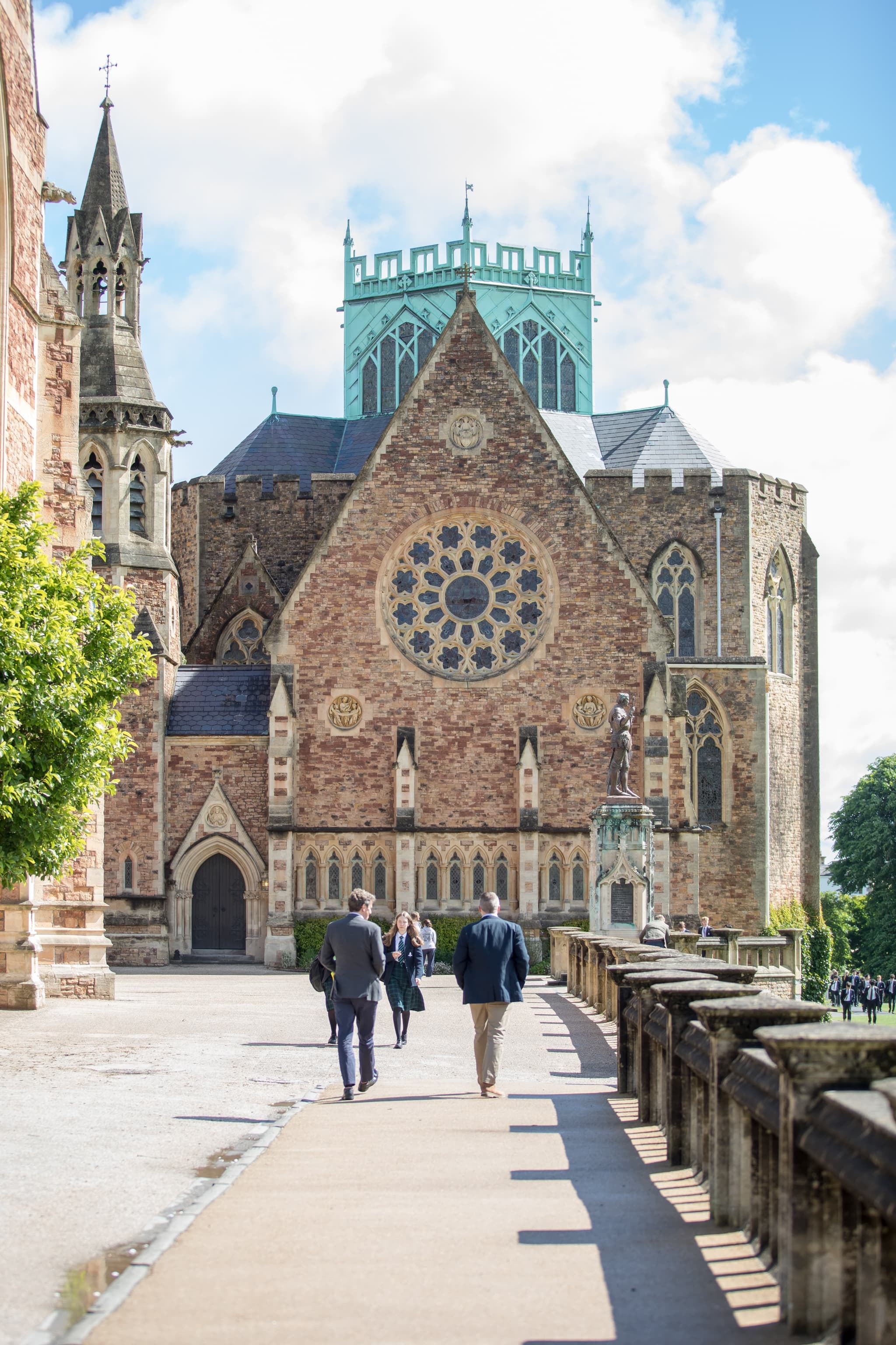 People walking towards a historic brick building with ornate architecture, a large rose window, and a green tower against a cloudy sky.