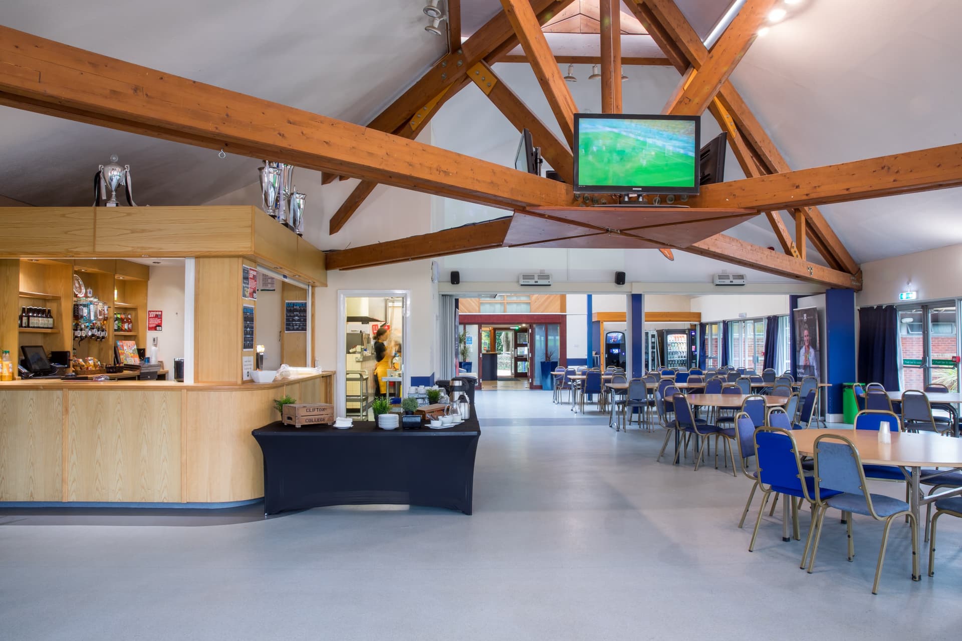 Spacious cafeteria with wooden beams, a bar area, tables and chairs, and a TV mounted on the ceiling showing a sports game.
