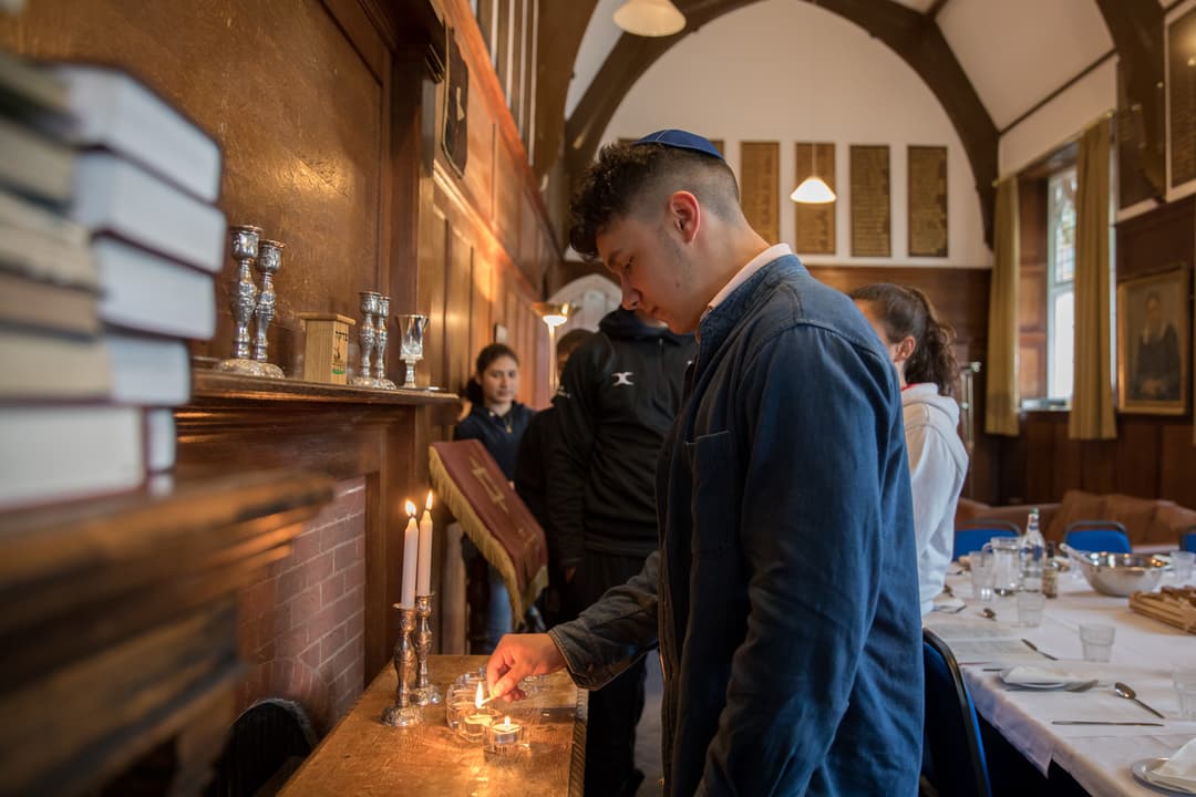 A person lights a candle in a room with wooden walls, books, and a set dining table, surrounded by others in a solemn setting.