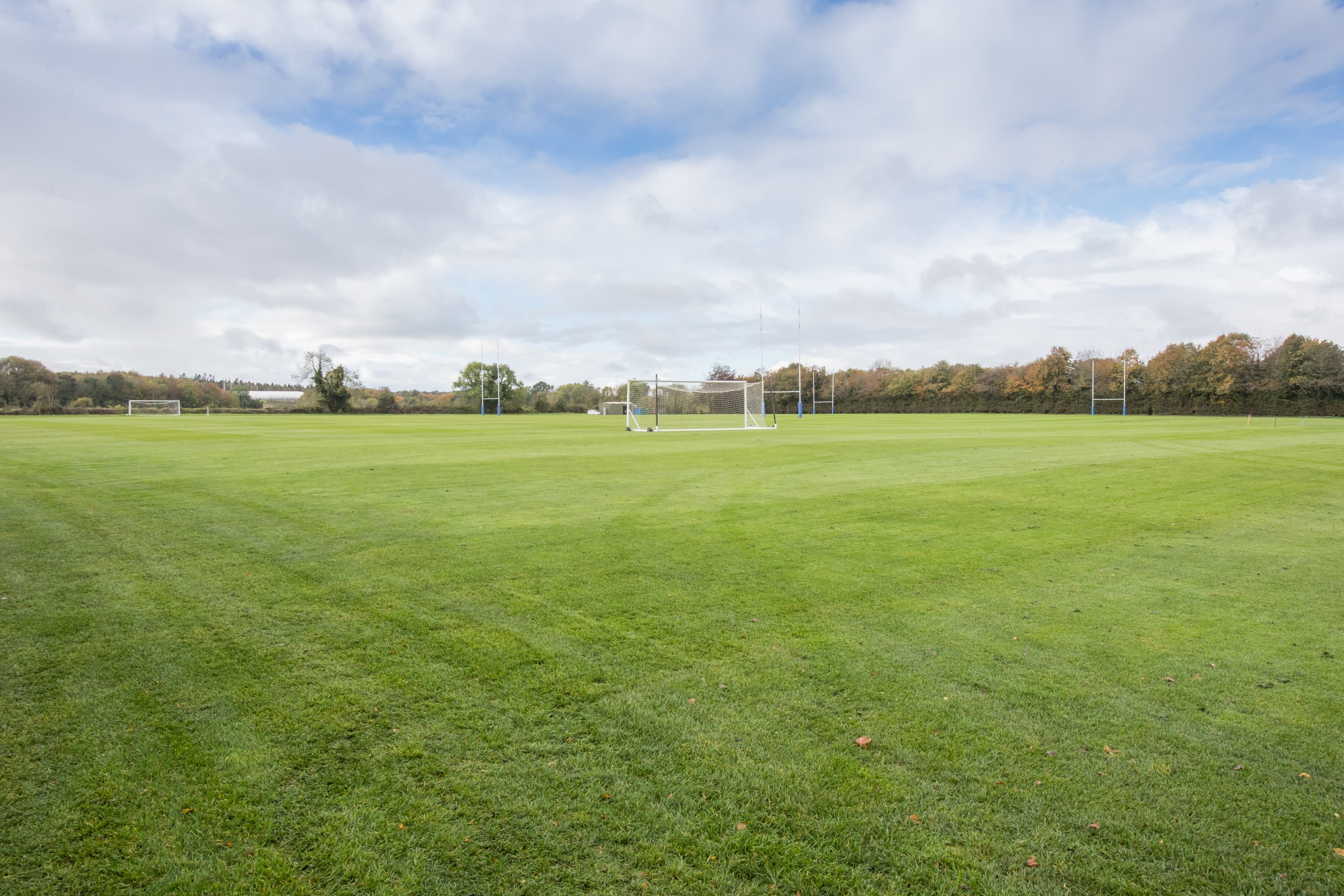 A vast, empty soccer field with green grass under a partly cloudy sky, surrounded by trees and distant goalposts.