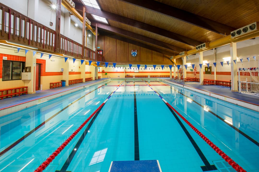 Indoor swimming pool with clear blue water, red lane dividers, and blue pennant flags. Wooden ceiling and benches line the sides.