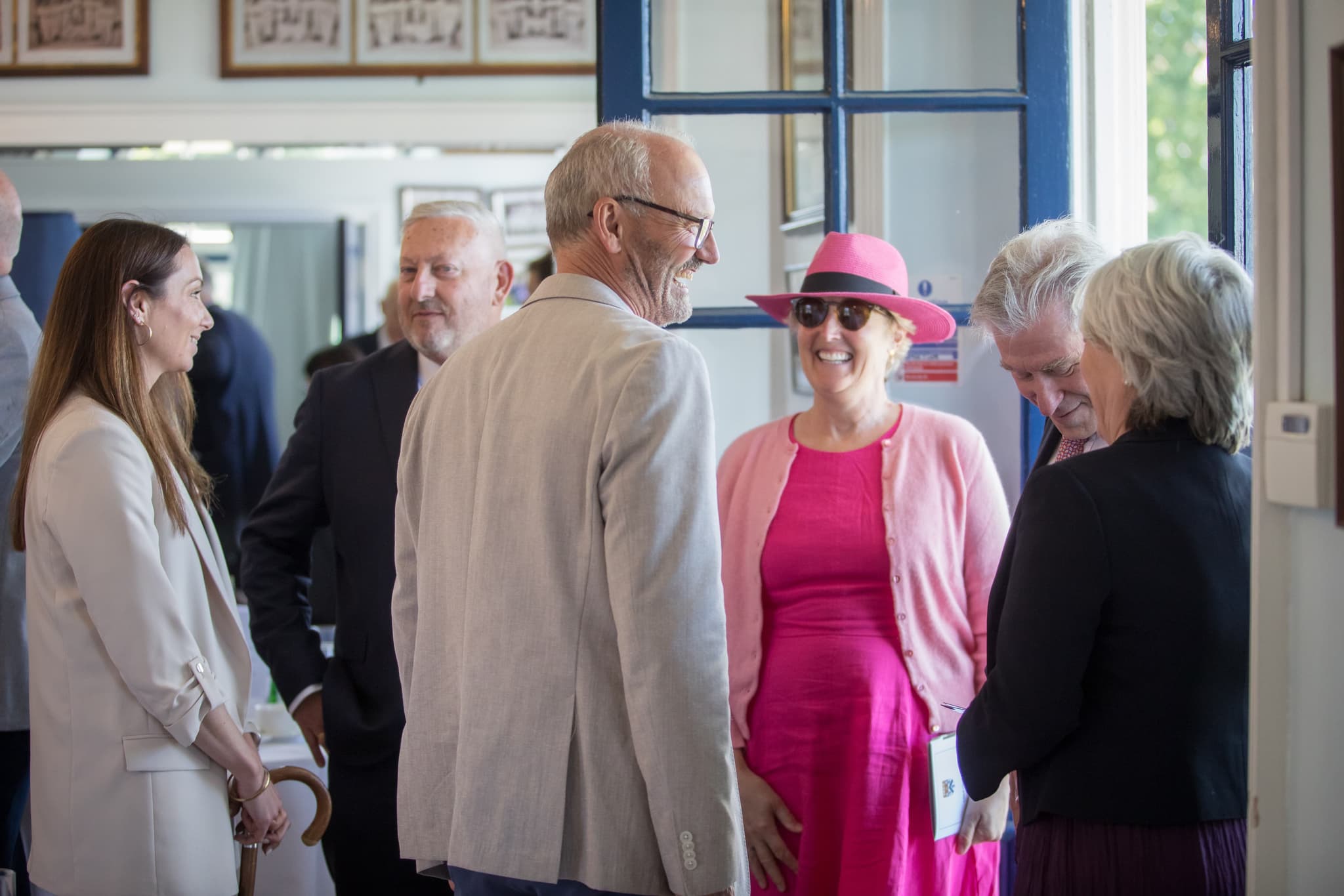 Group of people in formal attire chatting indoors; one woman wears a bright pink dress and hat.