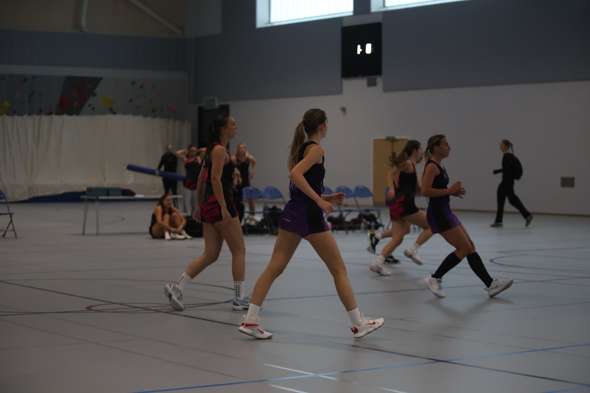 Indoor netball game in progress with players in action, wearing athletic gear. Spectators and gym equipment visible in the background.