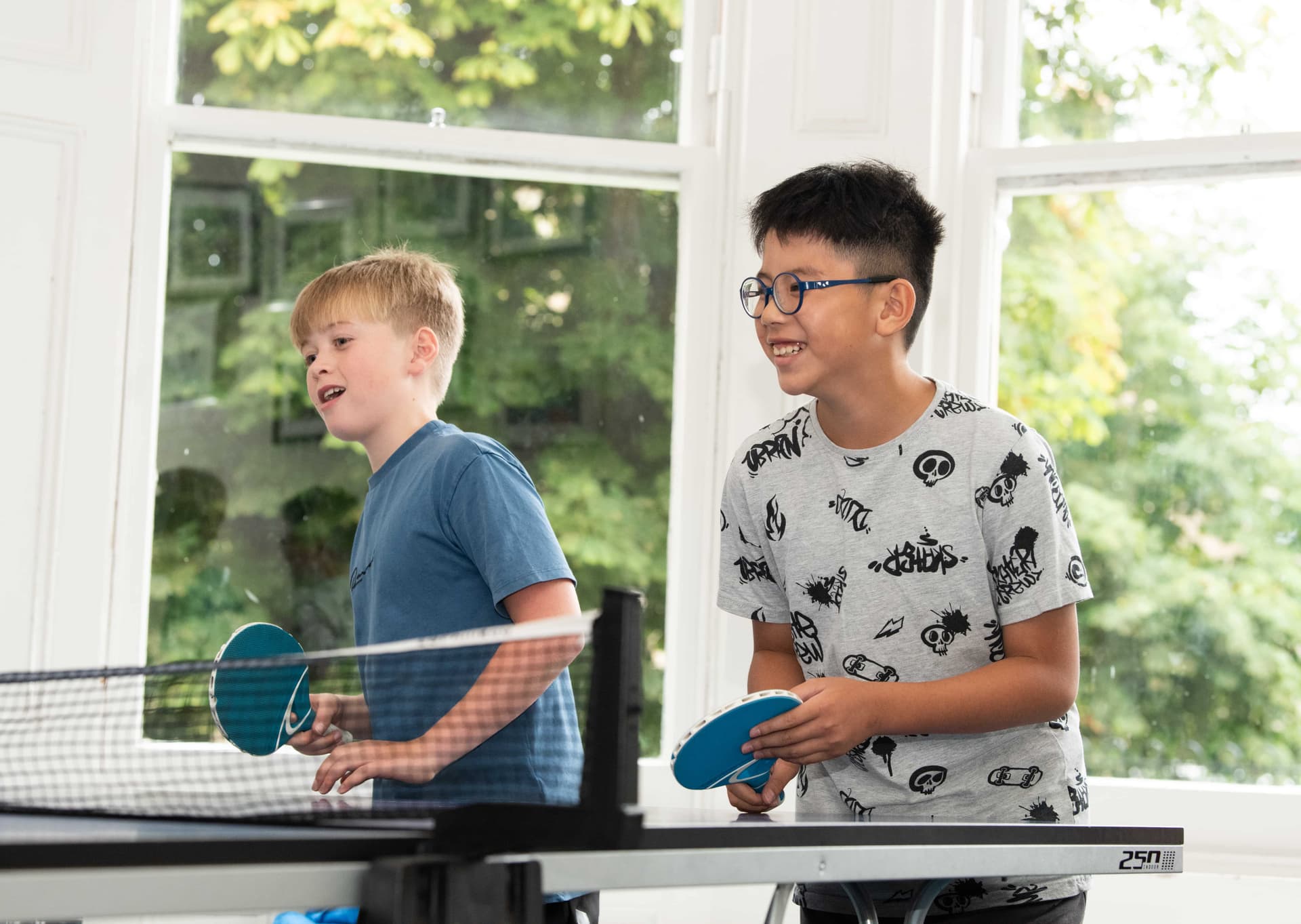 Two boys playing table tennis indoors, smiling and holding paddles, with large windows and greenery in the background.