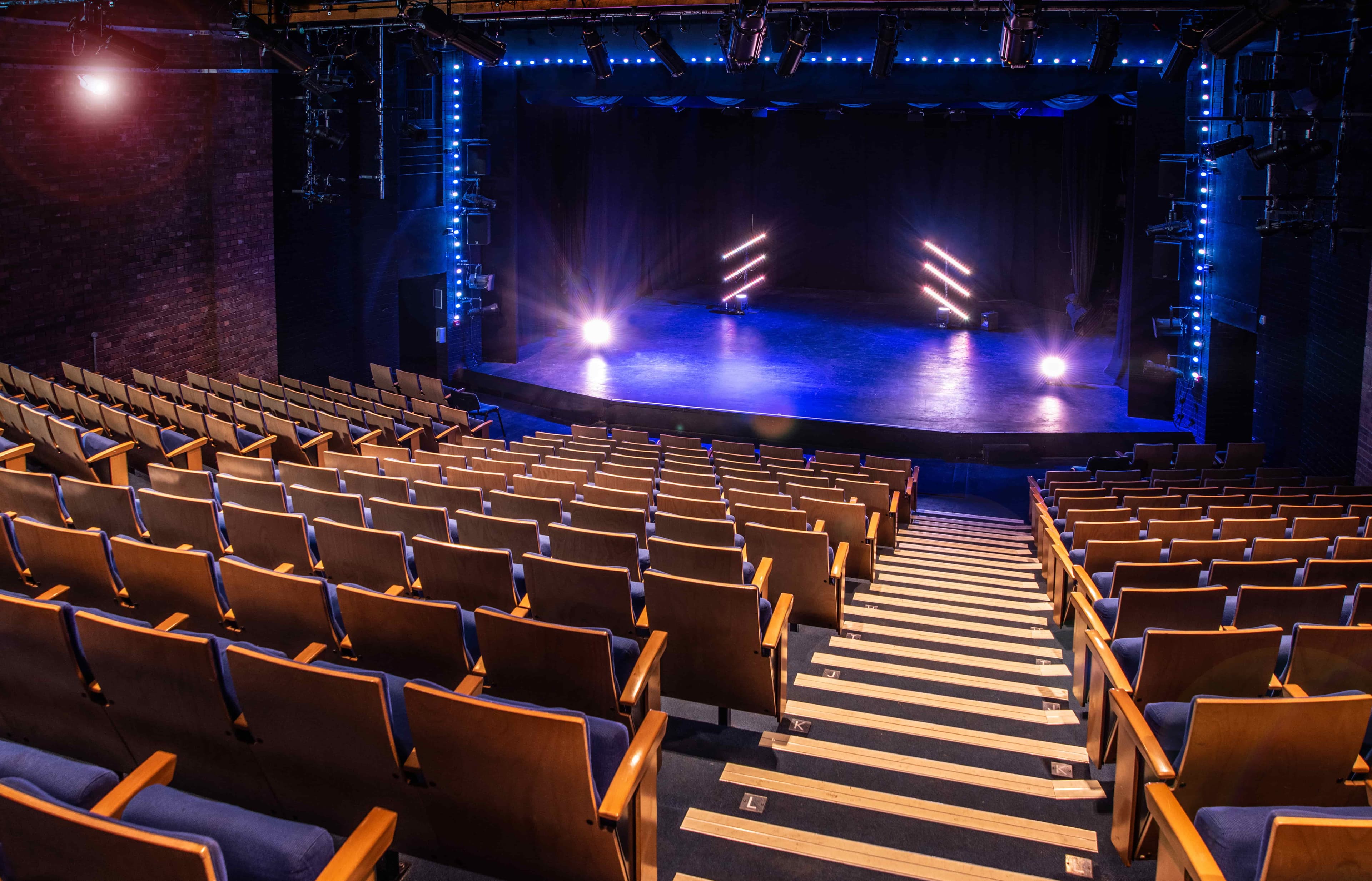 Empty theatre with rows of wooden seats facing a stage lit by blue and purple lights, creating a dramatic ambiance.