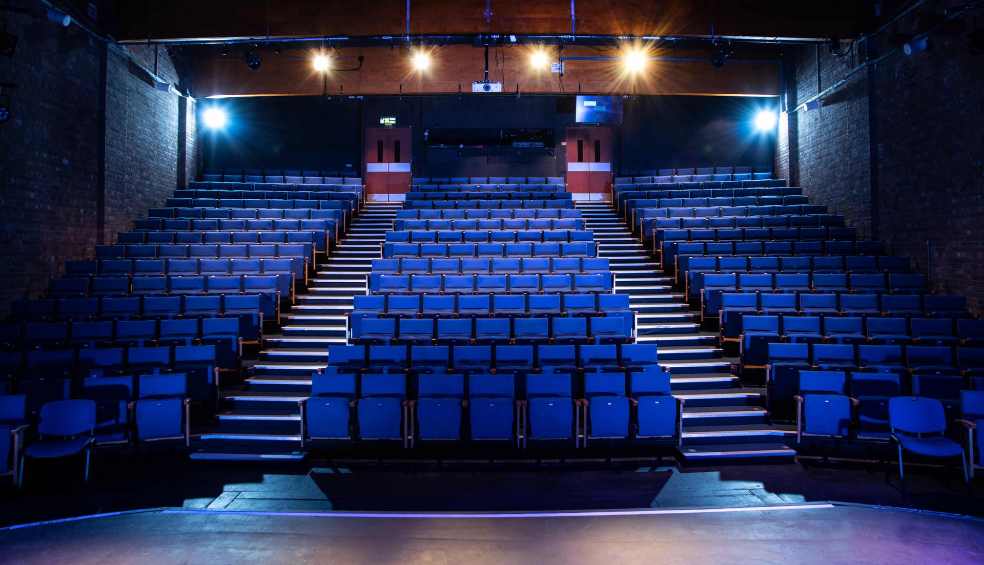 Empty theatre with rows of blue seats facing a stage, illuminated by bright overhead lights.