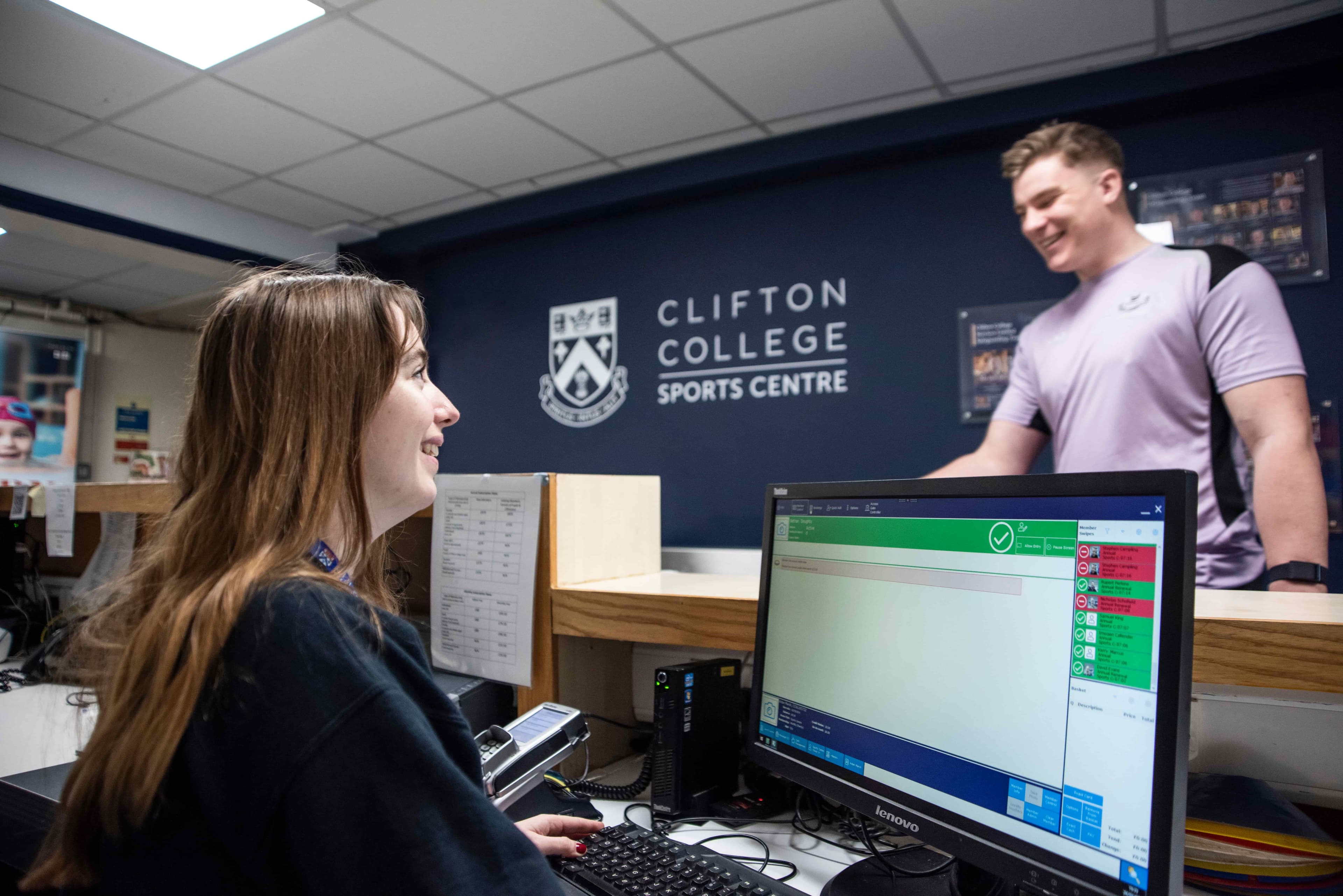 A woman at a computer greets a man standing at the Clifton College Sports Centre reception desk.