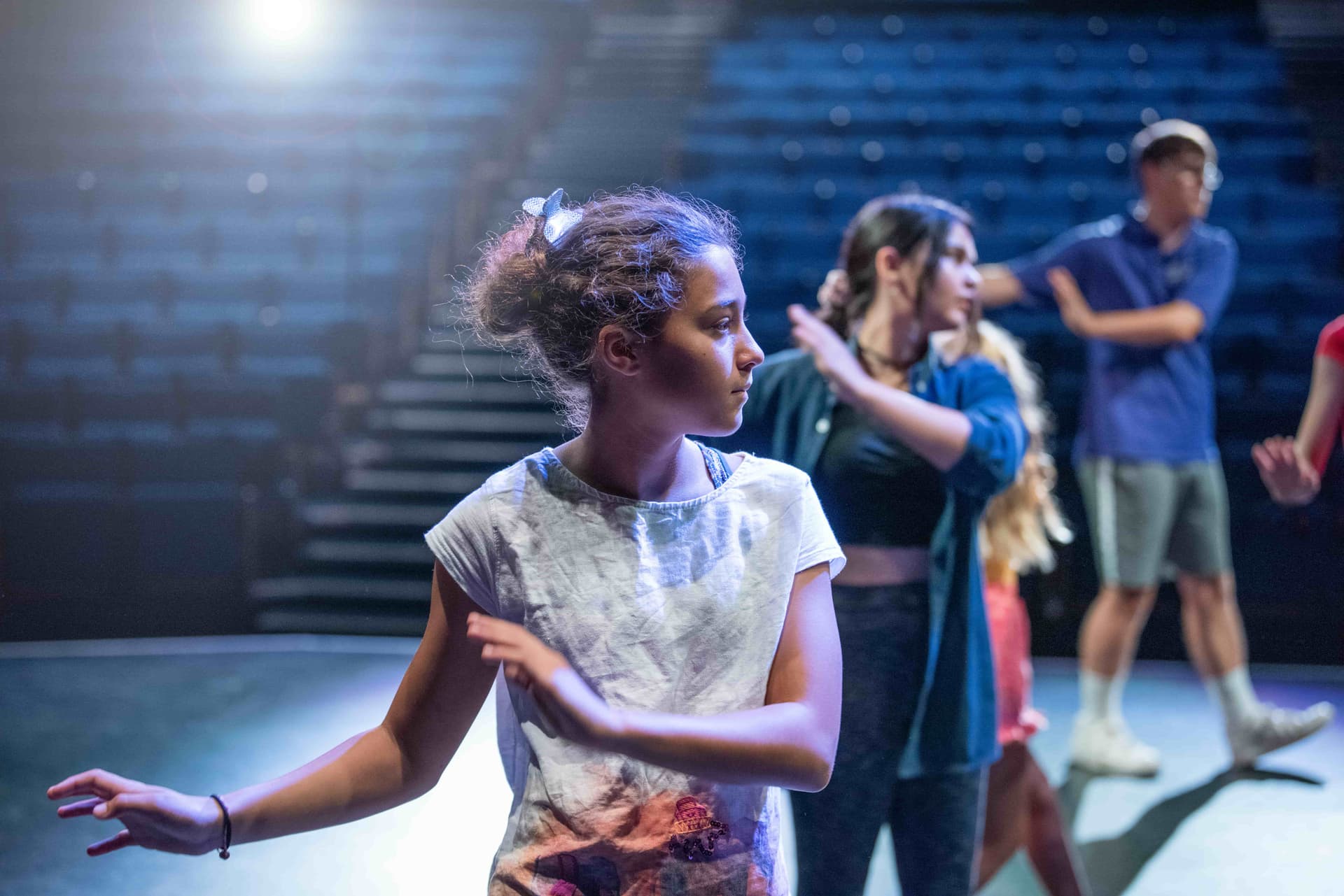 Young girl in a white shirt practicing dance on a dimly lit stage with other dancers in the background.