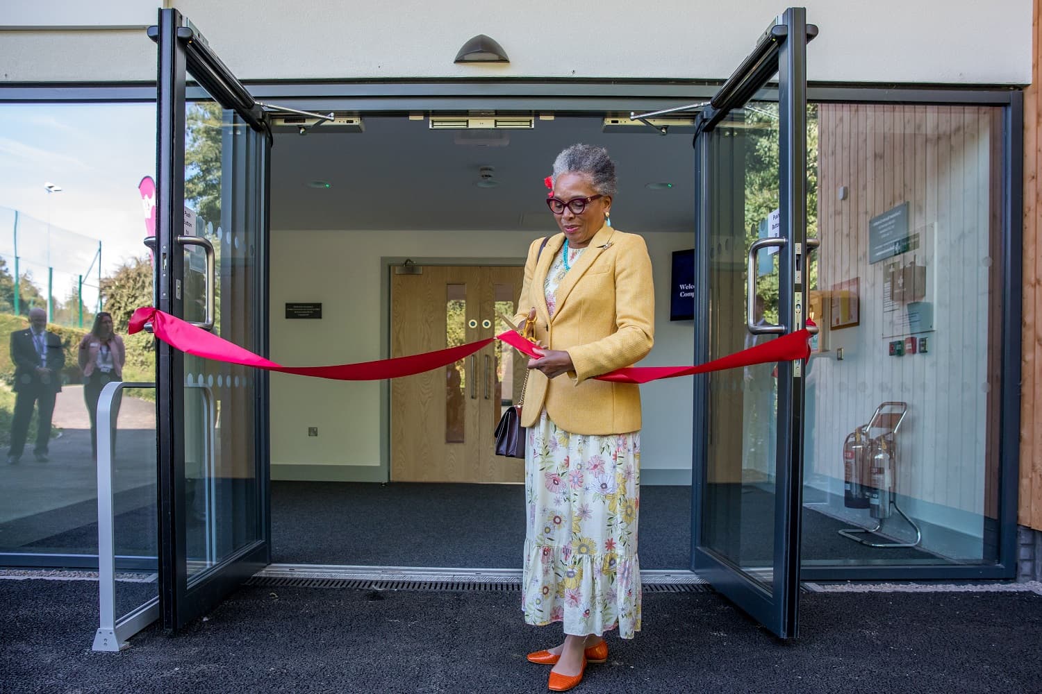 A woman in a yellow blazer and floral skirt cuts a red ribbon at a building entrance, signifying an opening ceremony.