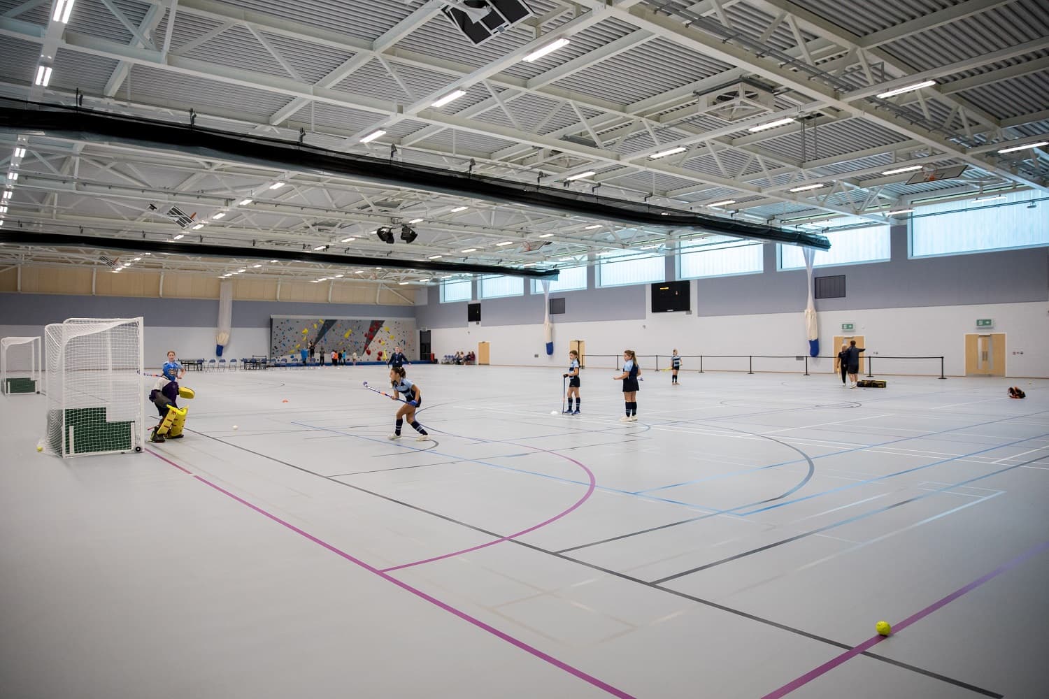 Indoor sports hall with people playing field hockey. Players wear uniforms, and a goalie stands near the net. Bright lighting and high ceiling.